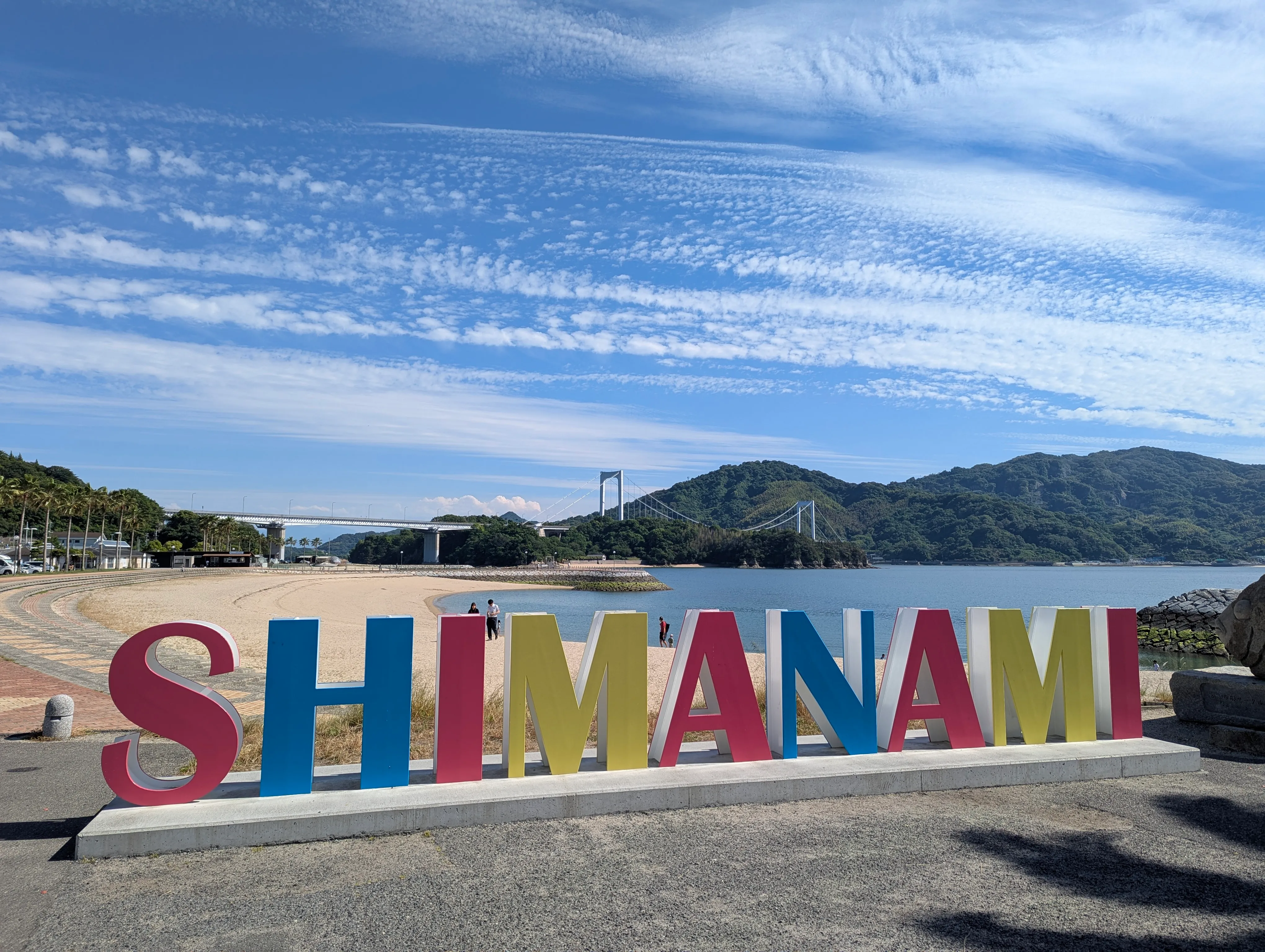 View of beach and suspension bridge with large colorful sign reading "Shimanami"
